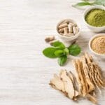 Flat lay of natural supplements including herbal capsules, medicinal roots, organic green powder, and botanical leaves arranged on a white wooden surface