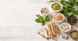 Flat lay of natural supplements including herbal capsules, medicinal roots, organic green powder, and botanical leaves arranged on a white wooden surface