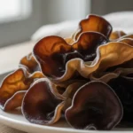 A close-up, editorial photograph showcases a cluster of fresh wood ear mushroom other names, highlighting their unique gelatinous texture and subtle earthy tones against a clean, neutral background with soft, natural lighting.
